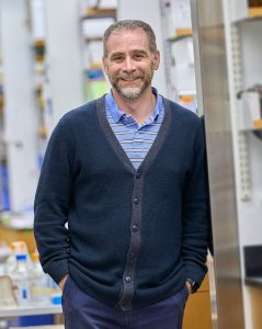 A man smiling while standing in a laboratory setting, wearing a navy blue cardigan over a light blue striped shirt. Shelves with laboratory equipment are visible in the background.