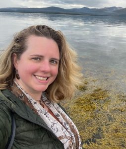 A woman with wavy hair smiles while standing near calm water, with yellow seaweed in the foreground and mountains in the background. The sky is overcast, suggesting a cool atmosphere.