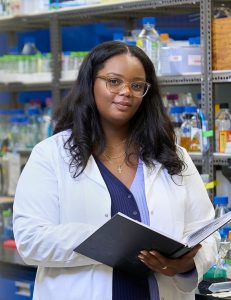 A woman wearing glasses and a lab coat stands in a laboratory filled with various bottles and containers. She holds a notebook and looks directly at the camera, with shelves of scientific equipment in the background.