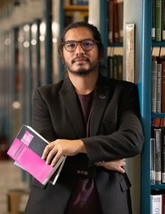 A person wearing glasses and a black blazer stands in a library, holding several books, including a pink one. Shelves filled with books are visible in the background.