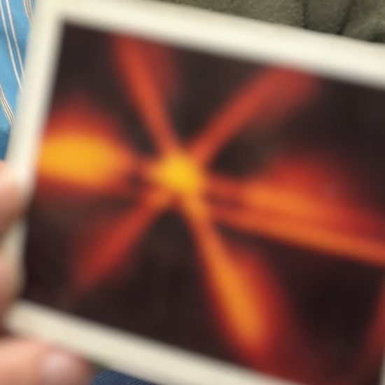 A person holding a blurry photograph showing a pattern of orange and yellow radiating lines against a dark background. The person is wearing a blue and white striped shirt.