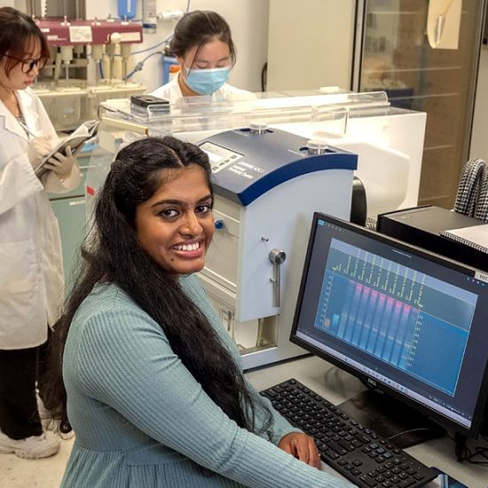 A woman with long dark hair smiles while working at a computer in a laboratory. Two other women in lab coats are in the background, one checking a device and the other holding a notebook. Laboratory equipment is visible.