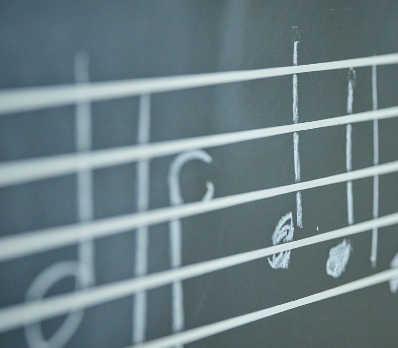Close-up of musical staff lines on a chalkboard, showing several notes written in white chalk. The background is dark, emphasizing the simplicity of the notes and lines.