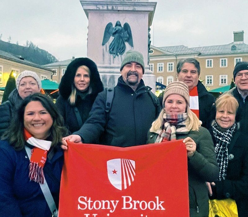 A group of eight individuals poses for a photo in front of a statue, holding a red banner with the Stony Brook University logo. They are dressed warmly, with some wearing scarves. The setting appears to be a square with buildings and trees in the background.