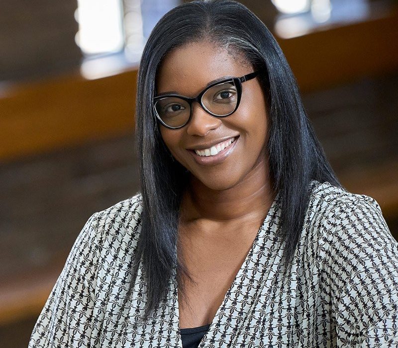 A woman with long, straight hair wearing glasses and a patterned black and white blazer smiles at the camera. She is positioned in a well-lit interior space with wooden accents in the background.