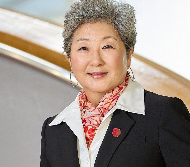 A woman with short gray hair wearing a black blazer and a white shirt, standing in front of a softly lit background. She has a pink and orange scarf around her neck and is smiling, showcasing a pin on her blazer.
