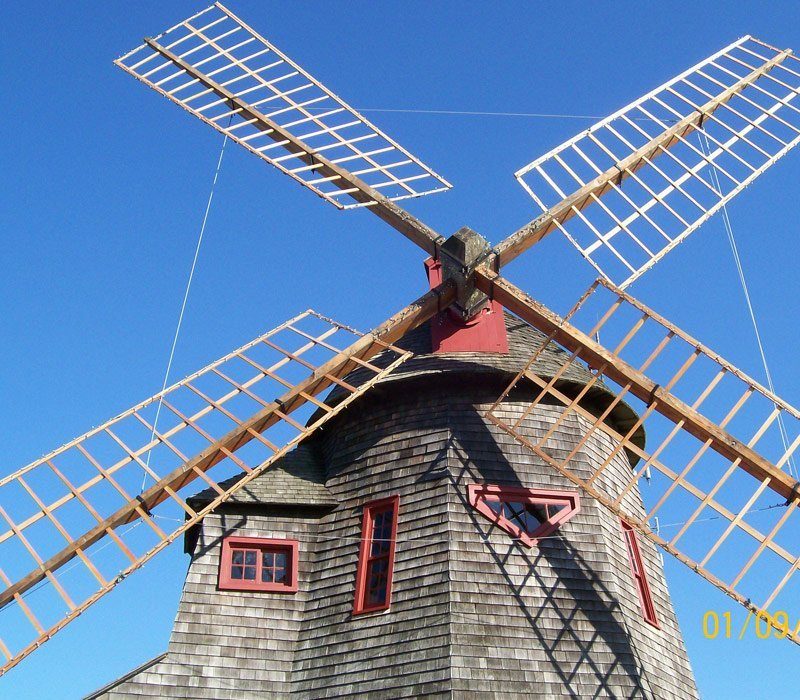 A traditional windmill with a wooden exterior and a conical roof, set against a clear blue sky. Its large, white sails are prominently displayed, and several red-tinted windows are visible on the structure's curved walls.