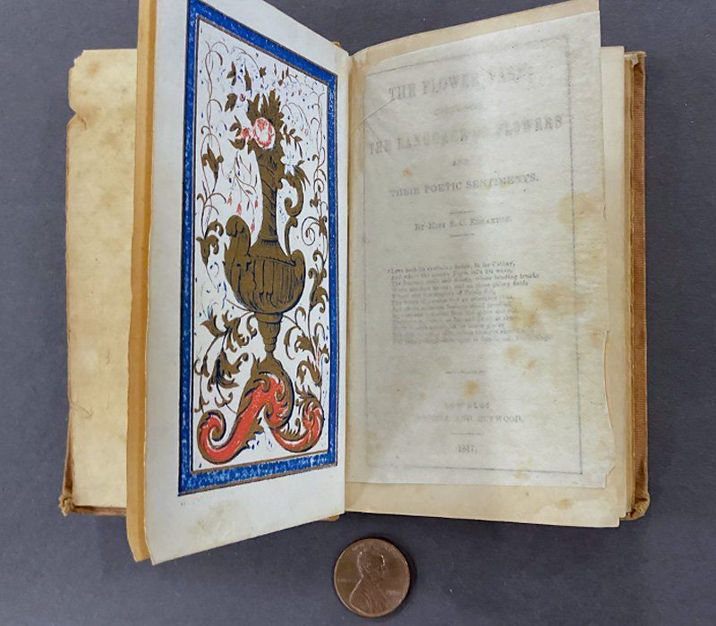An open antique book titled "The Flower Vase for Language of Flowers" featuring an intricately designed illustration with a bird and decorative elements. A small coin is placed on a dark surface beside the book.