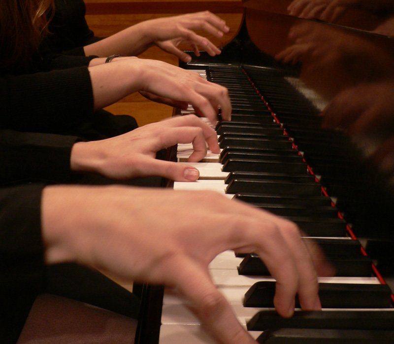 Close-up of multiple hands playing a piano. The fingers are positioned over the black and white keys, with some keys appearing pressed. The focus is on the motion of the hands, set against a blurred background.