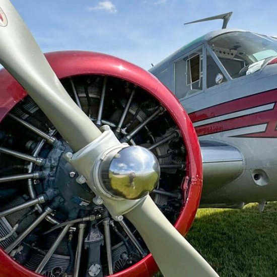 Close-up view of a vintage aircraft's propeller and engine. The propeller has a silver blade and a polished chrome hub. The engine, visible through the propeller, has several cylinders and is mounted on the aircraft, which features a red and silver color scheme.