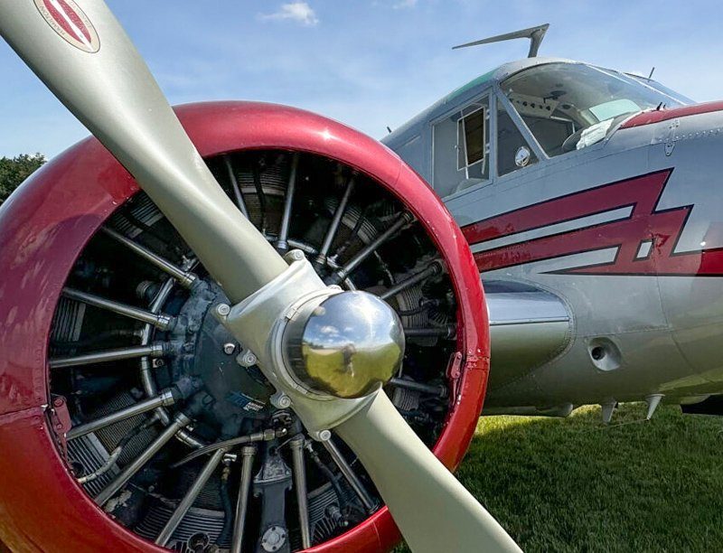 Close-up view of a vintage aircraft's propeller and engine. The propeller has a silver blade and a polished chrome hub. The engine, visible through the propeller, has several cylinders and is mounted on the aircraft, which features a red and silver color scheme.