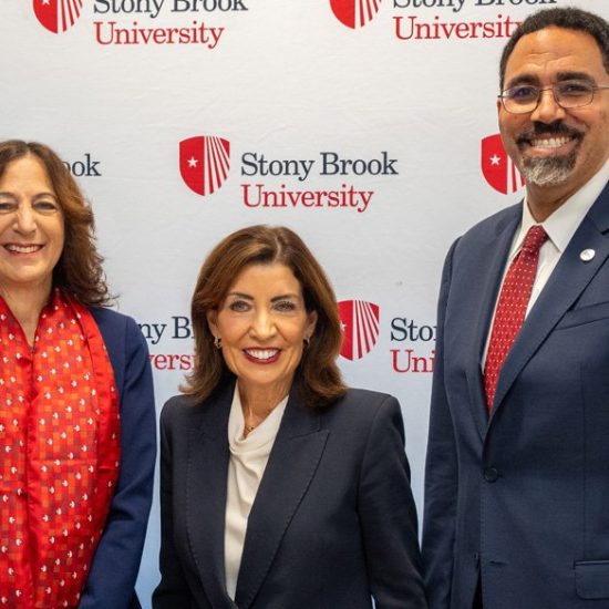 Three people pose for a photo at an event. On the left, a woman with curly hair and a red scarf; in the center, a woman wearing a dark suit; and on the right, a man in a navy suit with glasses. The background features a Stony Brook University banner.