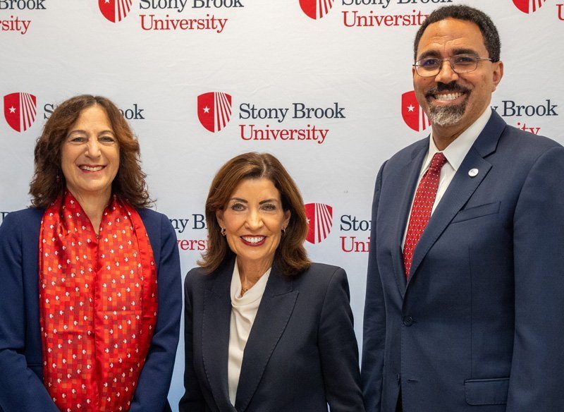Three people pose for a photo at an event. On the left, a woman with curly hair and a red scarf; in the center, a woman wearing a dark suit; and on the right, a man in a navy suit with glasses. The background features a Stony Brook University banner.
