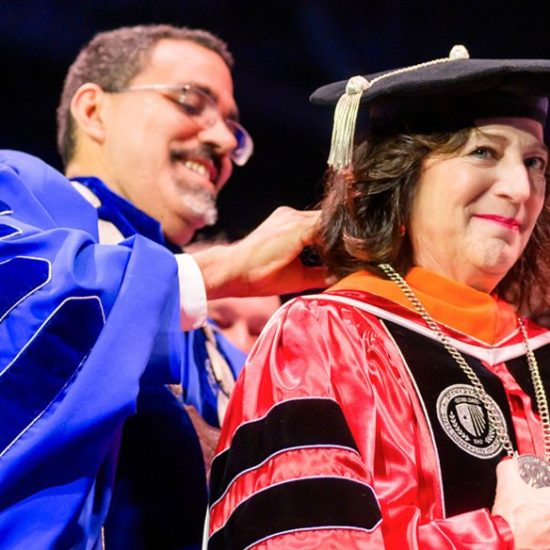 A man in a blue academic robe places a ceremonial hood on a woman wearing a red and black academic gown. The woman smiles as she receives the hood during a graduation ceremony, with a blurred audience in the background.