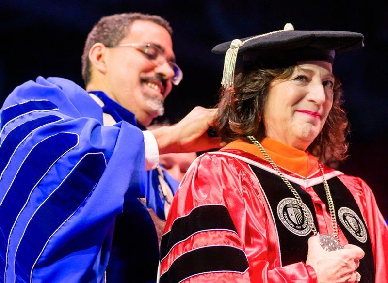 A man in a blue academic robe places a ceremonial hood on a woman wearing a red and black academic gown. The woman smiles as she receives the hood during a graduation ceremony, with a blurred audience in the background.