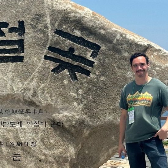 A man with a mustache stands next to a large stone with Korean characters etched on it. The backdrop features a coastal view of the ocean, and the man wears a green t-shirt and blue jeans, smiling at the camera.