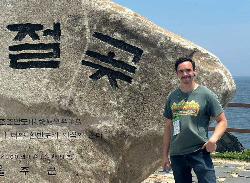 A man with a mustache stands next to a large stone with Korean characters etched on it. The backdrop features a coastal view of the ocean, and the man wears a green t-shirt and blue jeans, smiling at the camera.