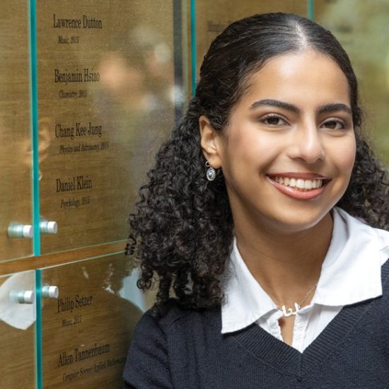 A smiling young woman with curly hair is standing next to a wall of plaques displaying names and years. She is wearing a dark sweater over a white collared shirt. Her earrings are visible, and there is a reflection in the glass behind her.