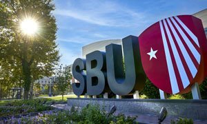 A large outdoor sign displaying the letters "SBU" next to a red shield with a white star and diagonal lines. The background features green trees and a blue sky, with sunlight filtering through the leaves.